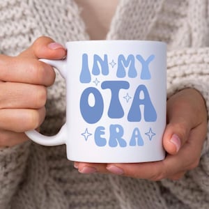 Könnte beinhalten: Weiße Keramiktasse mit dem Text "IN MY OTA ERA" in hellblauer Schrift im Retro-Stil. Die Tasse hat einen Standardgriff und wird von einer Person gehalten, die einen hellgrauen Pullover trägt.