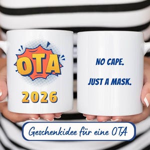 Könnte beinhalten: Zwei weiße Keramikbecher. Ein Becher zeigt den Text "OTA 2026" in Orange und Gelb mit einem blauen und weißen Burst-Design. Der andere Becher hat den Text "NO CAPE. JUST A MASK." in Blau. Die Becher werden von einer Person gehalten.