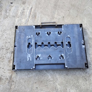 May include: A rectangular metal object with a handle, featuring cut-out shapes and slots. The object is a dark gray color and appears to be made of steel. The design includes oval and flame-shaped cutouts, and a handle for carrying.