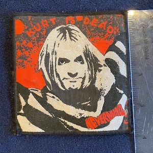 May include: Black and white embroidered patch with a red background. The patch features a portrait of Kurt Cobain with the text "Kurt is Dead" and "Nevermind" in red.