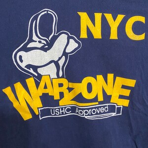 Puede incluir: Una camiseta azul marino con un gráfico amarillo de una mano de dibujos animados haciendo un signo de paz. El gráfico también incluye el texto "NYC WARZONE USHC Approved".