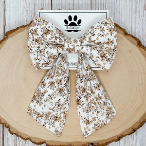 May include: A large, floral bow tie with a white background and brown flowers. The bow tie has long, flowing tails. The bow tie is on a wooden slice with the text "J&S PAWTIQUE" and a paw print logo.