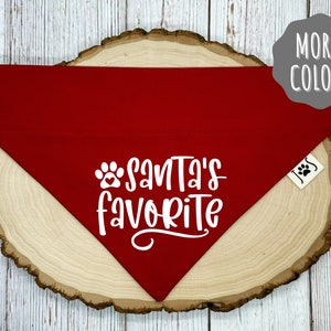 May include: A red dog bandana with the text "Santa's Favorite" in white script and a paw print. The bandana is displayed on a wooden slice, with a tag that says "More Colors".