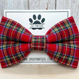 May include: Red and black plaid bow tie with yellow and blue accents. The bow tie is made of fabric and has a classic bow shape. The bow tie is on a white card with the text "J&S Pawtique" and "www.jandspawtique.com".