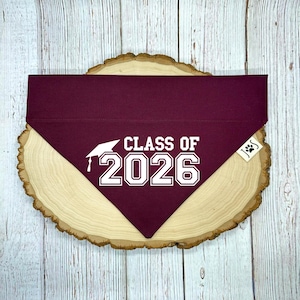 May include: A maroon dog bandana with the text "CLASS OF 2026" in white, along with a graduation cap graphic. The bandana is triangular and rests on a wooden slice with a light wood grain.