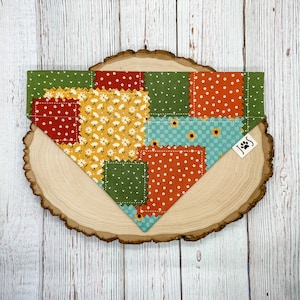 May include: A patchwork dog bandana with a variety of colors and patterns, including red, yellow, green, and blue. The bandana has a stitched edge and a small paw print logo in the corner.