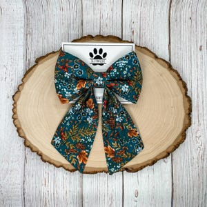 May include: A teal blue bow tie with a floral pattern featuring orange, white, and brown accents. The bow tie is made of fabric and has a classic bow shape.