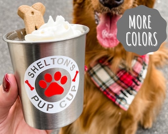 Può includere: Un bicchiere in acciaio inossidabile con un adesivo bianco che dice "Shelton's Pup Cup" con un'impronta di zampa rossa al centro. Il bicchiere è guarnito con panna montata e un dolcetto per cani. Sullo sfondo c'è un golden retriever che indossa un bandana a quadri rossi e bianchi.