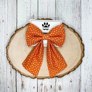 May include: An orange fabric bow with a white polka dot pattern, featuring two long, pointed tails hanging downward. The bow is attached to a white rectangular display card that displays a black paw print logo and the text J&S PAWTIQUE.
