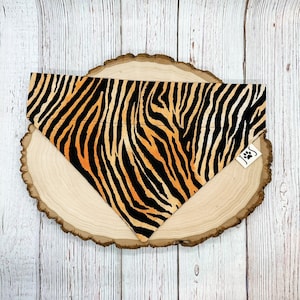 May include: Orange and black tiger print dog bandana. The bandana is on a wooden surface.
