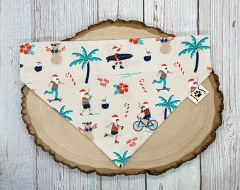May include: A white dog bandana with a Christmas-themed print featuring Santa Claus surfing, skateboarding, and riding a bicycle. The bandana has a tropical theme with palm trees, pineapples, and candy canes. The text "Ho Ho Ho" is printed on the bandana.