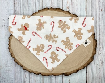 May include: White cotton dog bandana with a gingerbread man and candy cane pattern. The bandana has a red and white candy cane pattern and gingerbread men in various poses. The bandana has a small white tag with the text "JJS" on it.