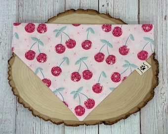 May include: Pink dog bandana with a cherry print. The bandana has a small tag with the letters "JJS" and a paw print.