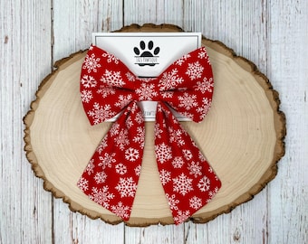 May include: A red fabric bow tie with a white snowflake pattern. The bow tie is displayed on a wooden slice with a white card that has a paw print logo and the text "J&S PAWTIQUE".