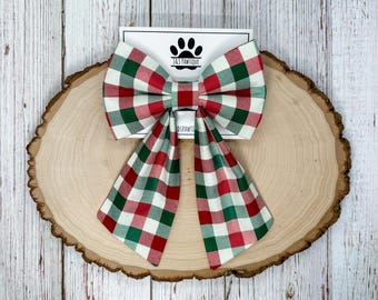 May include: A large, festive dog bow tie with a red, green, and white plaid pattern. The bow tie is displayed on a wooden slice with a white card featuring a paw print logo and the text "J&S PAWTIQUE".