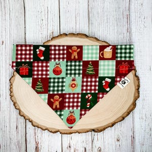 May include: A Christmas-themed dog bandana featuring a patchwork design with festive elements. The bandana showcases red and green squares with images of gingerbread men, ornaments, Christmas trees, and gift boxes. The bandana is displayed on a wooden slice.