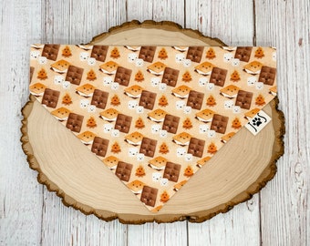 May include: A tan dog bandana with a pattern of brown chocolate bars, white marshmallows, and orange flames. The bandana has a small brown tag with the letter "J" on it.