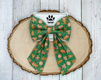 May include: A green and brown bow with a gingerbread man and candy cane print. The bow is tied with a white ribbon and has a small tag that says "J&S Pawtique".