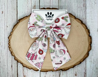 May include: A festive pink and white dog bow tie with a Christmas-themed pattern. The bow tie features Santa Claus, Christmas trees, ornaments, and candy canes. The bow tie is displayed on a wooden slice with the text "JAS PAWTIQUE".
