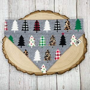 May include: A gray bandana with a Christmas tree pattern. The trees are in various colors and patterns, including red and black plaid, green and white checkered, leopard print, and solid white. The bandana has a small tag with the text "J&S" on it.