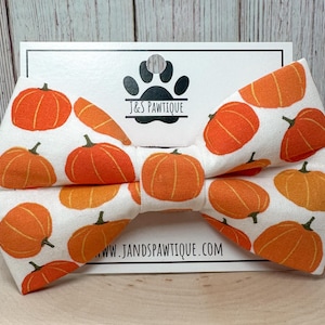 May include: A white bow tie with a repeating pattern of orange pumpkins. The bow tie is on a white card with the text "J&S PAWTIQUE.COM" and a paw print logo. A festive accessory for pets.