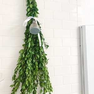 May include: A bunch of green leafy branches hanging from a white tiled wall with a shower head attached to the branches with a white ribbon.