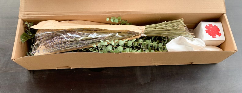 May include: A cardboard box filled with dried lavender and eucalyptus stems, a white cloth bag, and a small white box with a red cross.