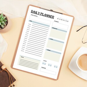 Könnte beinhalten: Ein Holz-Klemmbrett mit dem Aufdruck "DAILY PLANNER", mit Abschnitten für Aufgaben, Erinnerungen und Mahlzeiten. Eine kleine Sukkulente in einem braunen Topf, eine Tasse Kaffee und eine Brille befinden sich ebenfalls auf der hellgelben Oberfläche.