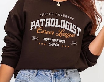 Puede incluir: Una sudadera marrón oscuro con el texto "SPEECH LANGUAGE PATHOLOGIST Career League" en letras blancas y naranjas. La sudadera también presenta el texto "MORE THAN JUST SPEECH" y el año "1900".