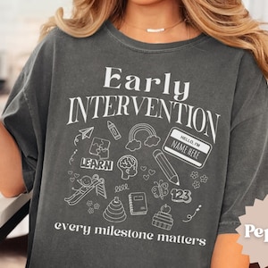 Peut inclure: T-shirt gris foncé avec l'inscription blanche "Early Intervention" et "every milestone matters". Le t-shirt présente des illustrations blanches de symboles éducatifs, comme un arc-en-ciel et un crayon.