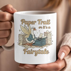 May include: White ceramic mug featuring a whimsical illustration. A goose wearing glasses and a blazer types on a vintage typewriter, surrounded by stacks of paper. The text reads "Paper Trail or it's a Fairytale" in a decorative font.
