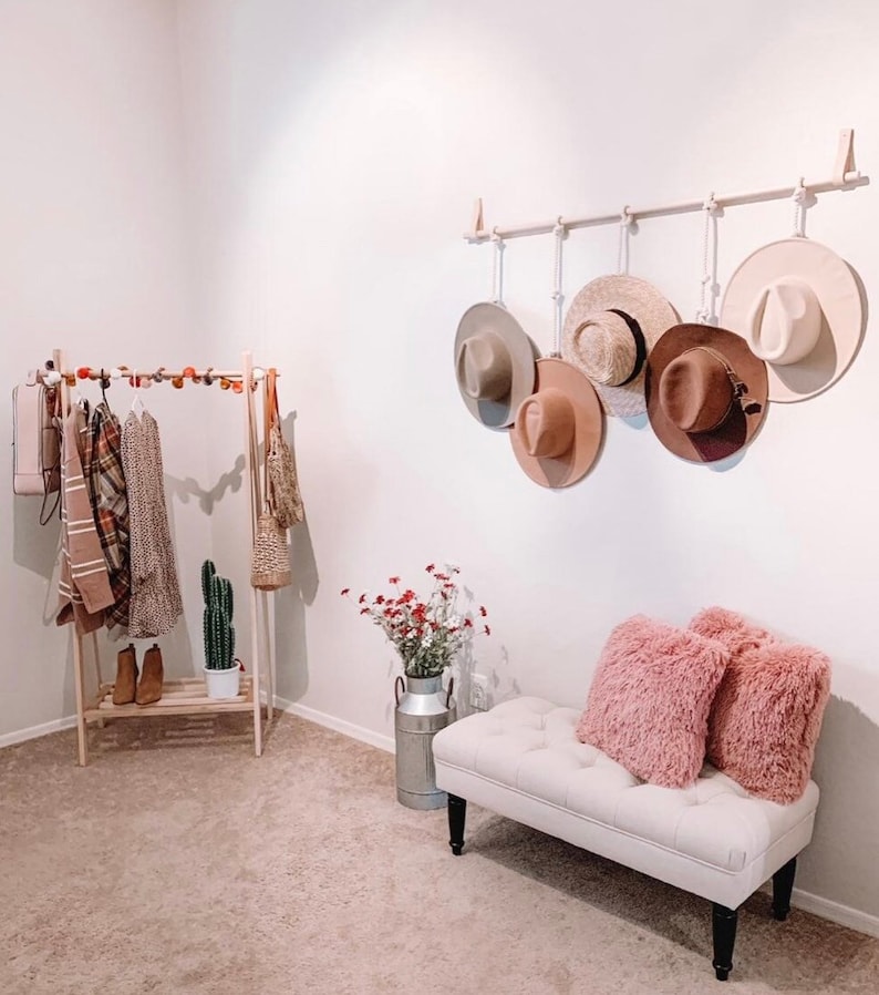 Pode incluir: Um quarto iluminado com um cabide de madeira exibindo v&aacute;rias roupas e acess&oacute;rios. Um chap&eacute;u de parede cont&eacute;m v&aacute;rios chap&eacute;us. Um banco com almofadas rosa e um vaso decorativo com flores completam a cena, criando um espa&ccedil;o elegante e organizado.