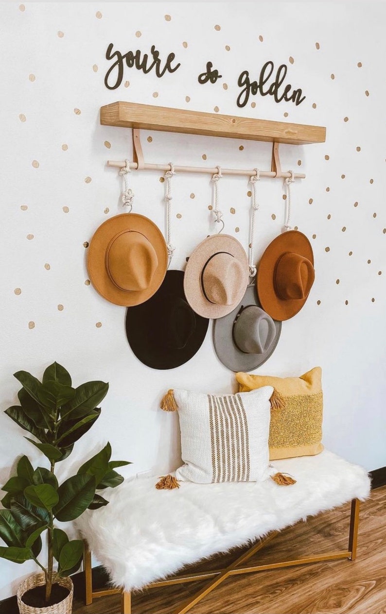 Pode incluir: Um cabide de parede com seis chap&eacute;us fedora em v&aacute;rias cores, incluindo castanho, preto e cinzento. O cabide &eacute; feito de madeira e corda, com as palavras "you're so golden" acima. Um banco com almofadas e uma planta completam a cena.
