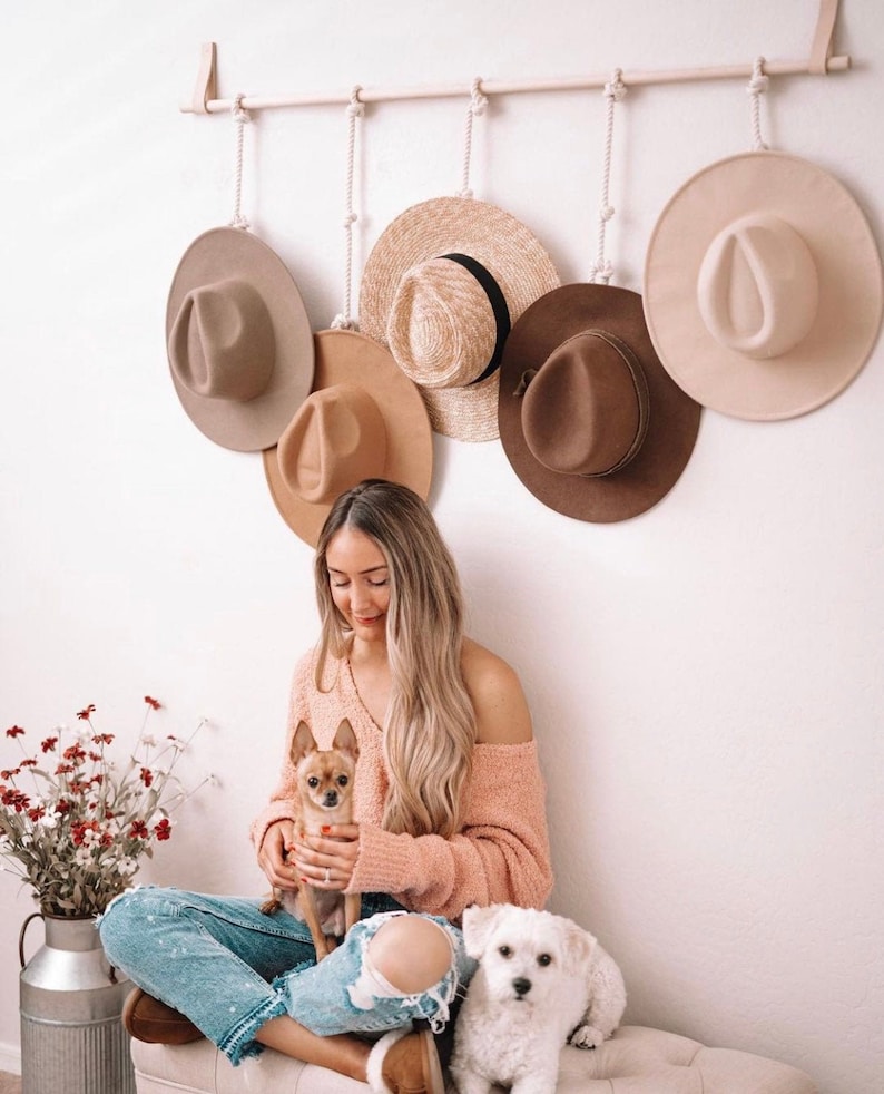Pode incluir: Uma cole&ccedil;&atilde;o de chap&eacute;us em tons de castanho e bege est&aacute; pendurada numa prateleira de madeira. Os chap&eacute;us est&atilde;o suspensos por cordas. Uma mulher est&aacute; sentada num banco com dois c&atilde;es pequenos. A mulher usa um su&eacute;ter rosa e jeans azuis.