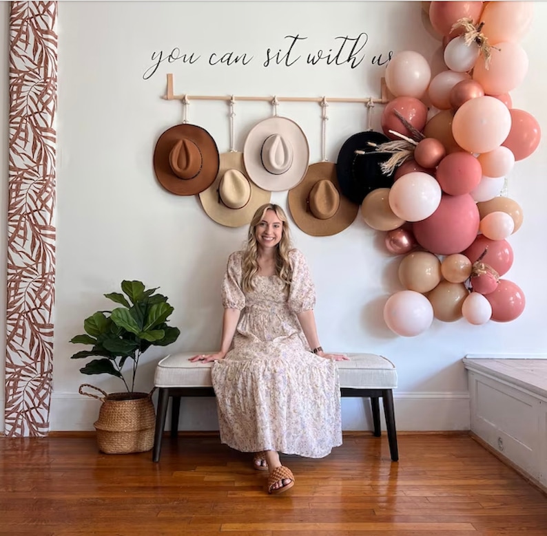 Pode incluir: Uma mulher senta-se num banco em frente a uma parede decorada com chap&eacute;us e bal&otilde;es. A parede tem o texto "you can sit with us". Uma variedade de chap&eacute;us de diferentes cores penduram-se numa prateleira de madeira. Uma planta numa cesta de vime est&aacute; &agrave; esquerda.