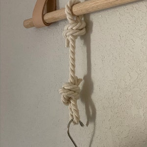 Pode incluir: Um suporte de madeira natural com uma corda creme pendurada. A corda &eacute; amarrada em dois lugares e tem um gancho em forma de S de metal na parte inferior. Uma tira de couro est&aacute; presa ao suporte.
