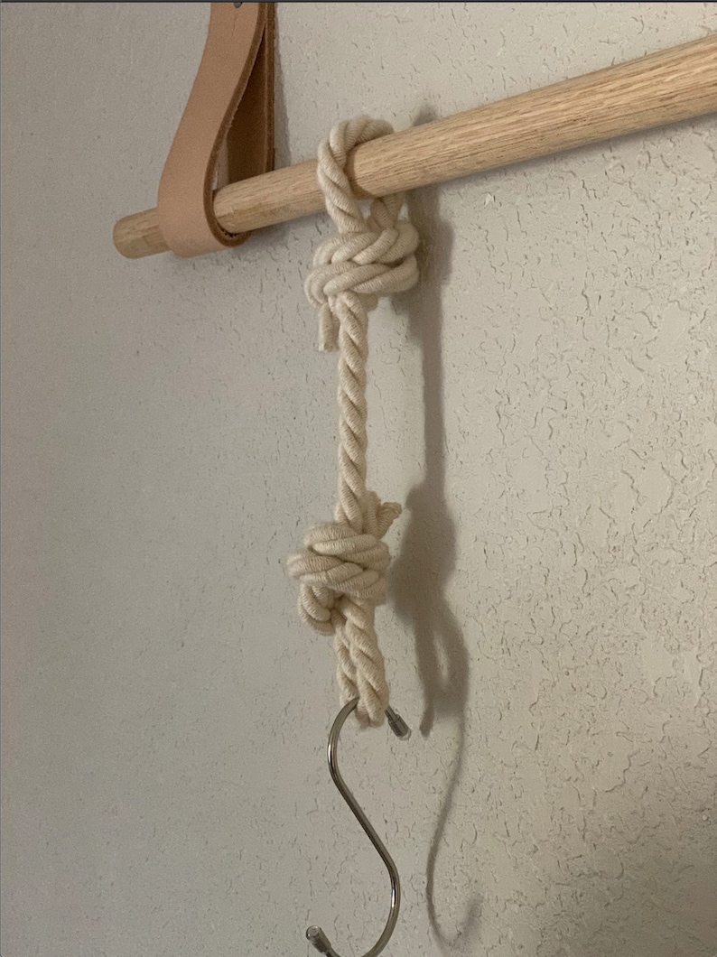 Pode incluir: Um suporte de madeira natural com uma corda creme pendurada. A corda &eacute; amarrada em dois lugares e tem um gancho em forma de S de metal na parte inferior. Uma tira de couro est&aacute; presa ao suporte.