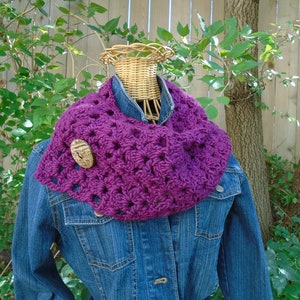 May include: Purple crocheted infinity scarf with a wooden button accent. The scarf is displayed on a denim jacket.