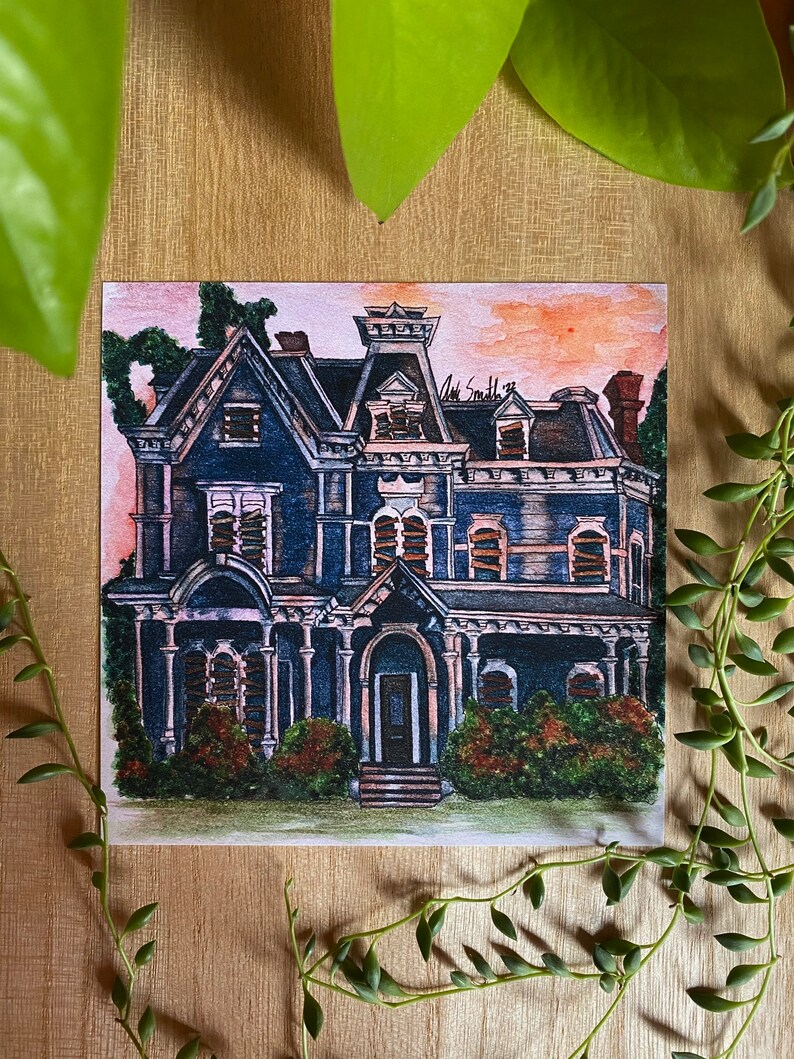 K&ouml;nnte beinhalten: Aquarellmalerei eines dunkelblauen viktorianischen Hauses mit schwarzem Dach und wei&szlig;em Zierrat. Das Haus hat mehrere Fenster und eine Veranda. Der Himmel ist eine Mischung aus Rosa und Orange. Gr&uuml;nes Laub umgibt das Haus. Die Signatur des K&uuml;nstlers befindet sich in der oberen rechten Ecke.