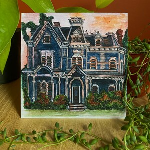 K&ouml;nnte beinhalten: Quadratischer Kunstdruck eines detaillierten blauen viktorianischen Hauses mit dunklem Dach und wei&szlig;en Zierleisten. Das Kunstwerk zeigt eine Veranda, mehrere Fenster und viel Gr&uuml;n. Die Signatur des K&uuml;nstlers, "Art Smith '22", ist oben rechts sichtbar.