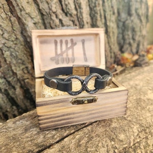 May include: A black leather bracelet with a silver double circle design. The bracelet is in a wooden box with a carved inscription.
