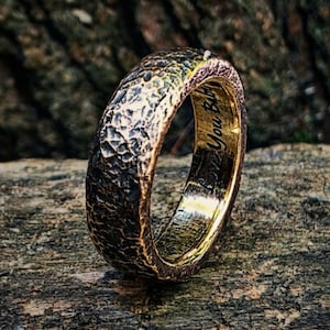 May include: A ring with a dark, heavily textured, hammered exterior surface and a smooth, polished gold-toned interior. The inner band features the engraved text "Love You Baby" in a cursive script. The ring has a thick, rounded profile.