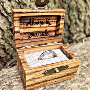 May include: Open wooden ring box with a silver ring inside, set against a natural background. The box is engraved with the words "Always by your side." The box has a brass clasp and hinges.