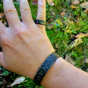 May include: A dark gray metal ring and cuff bracelet set. The ring is on a finger, and the bracelet is on a wrist. Both pieces have a textured, hammered appearance, with a dark, almost black finish. The background is blurred green grass and leaves.