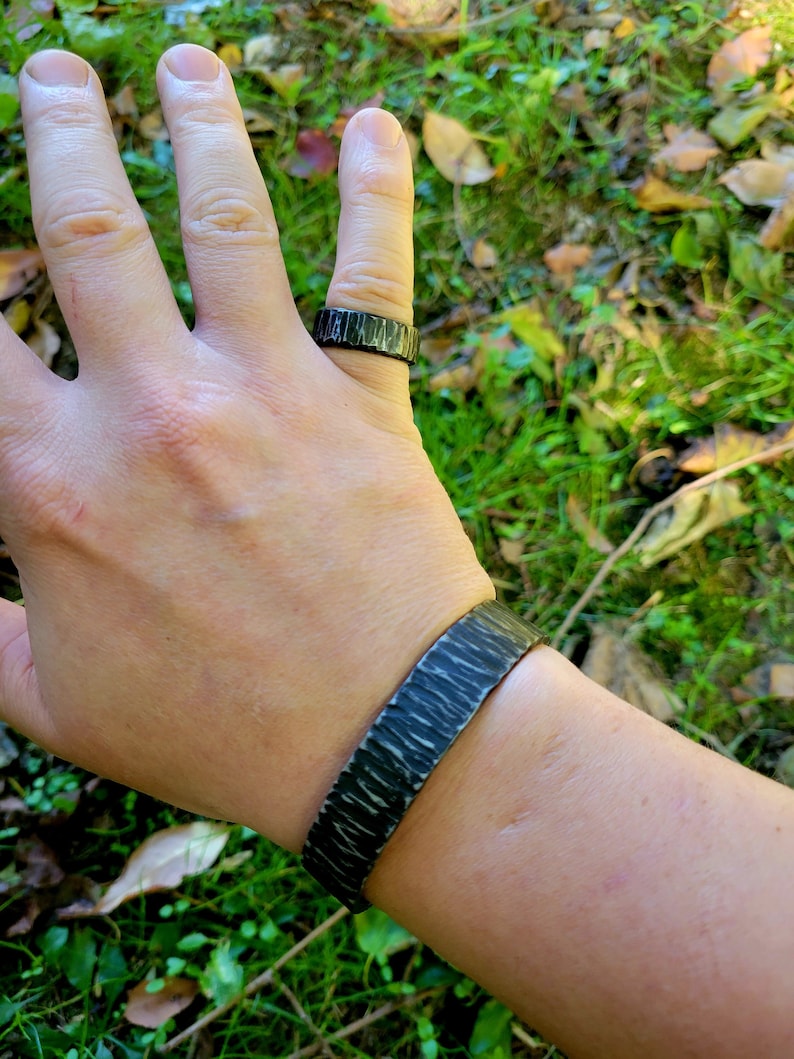 May include: A dark gray metal ring and cuff bracelet set. The ring is on a finger, and the bracelet is on a wrist. Both pieces have a textured, hammered appearance, with a dark, almost black finish. The background is blurred green grass and leaves.