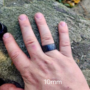 May include: A black ring with a textured surface is worn on a finger. The ring is 10mm wide. The ring's design features a pattern of vertical lines. The ring is on a hand.