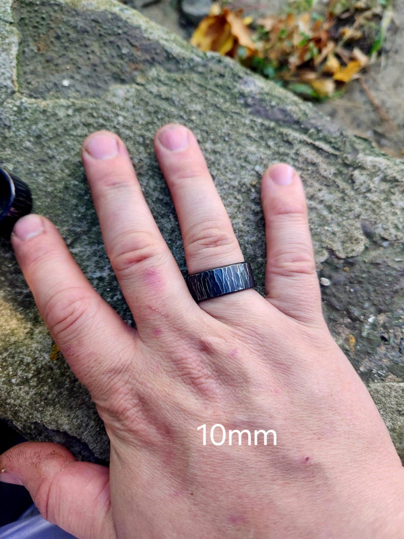 May include: A black ring with a textured surface is worn on a finger. The ring is 10mm wide. The ring's design features a pattern of vertical lines. The ring is on a hand.