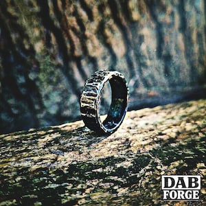 A textured, dark gray ring with a hammered metal appearance. The ring is set against a blurred natural background, highlighting its rugged, handcrafted aesthetic. The ring's surface has a unique, irregular pattern.