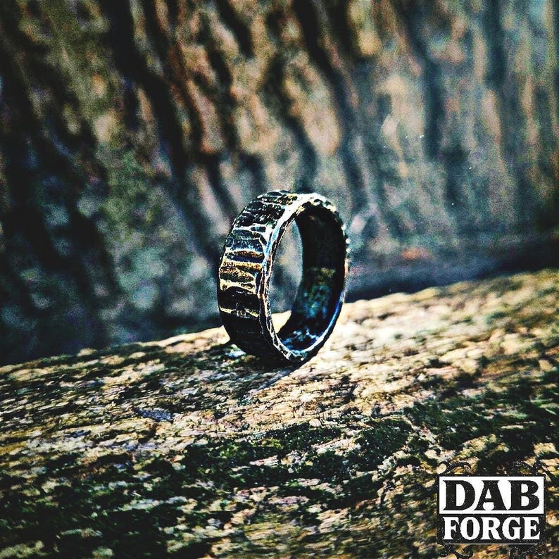 A textured, dark gray ring with a hammered metal appearance. The ring is set against a blurred natural background, highlighting its rugged, handcrafted aesthetic. The ring's surface has a unique, irregular pattern.
