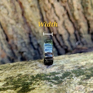 May include: A dark, textured ring with a hammered metal appearance. The ring is set against a natural wood background. The word "Width" is written in yellow above the ring.